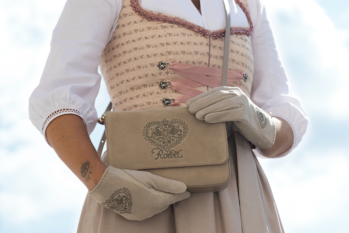 A woman in a dirndl is carrying a light leather bag adorned with traditional embroidery, along with light leather gloves featuring heart designs.