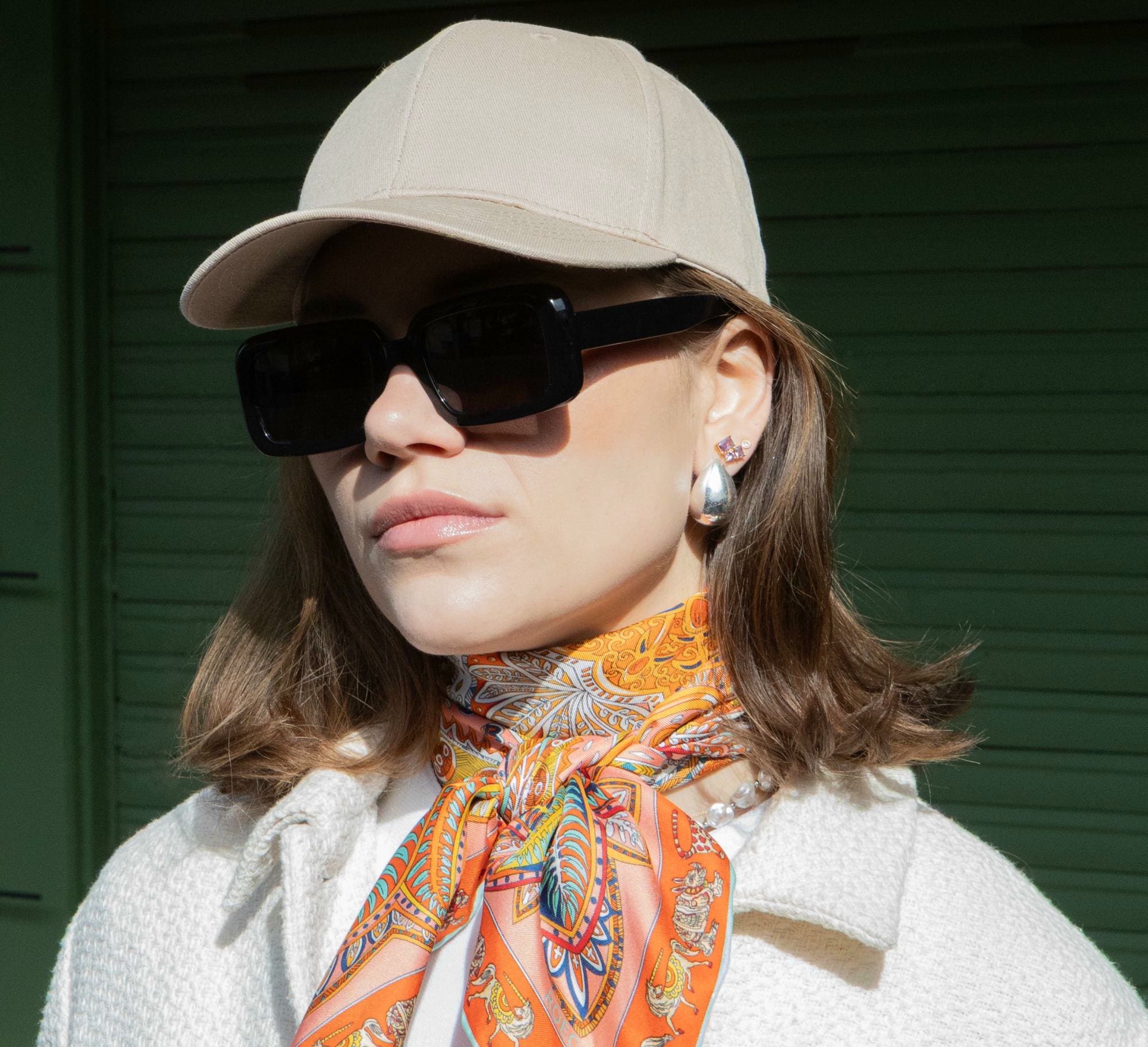 Eine junge Dame mit schulterlangem Haar trägt eine helle Baseball-Cap und einem orangenem Seidentuch um den Halt gebunden.