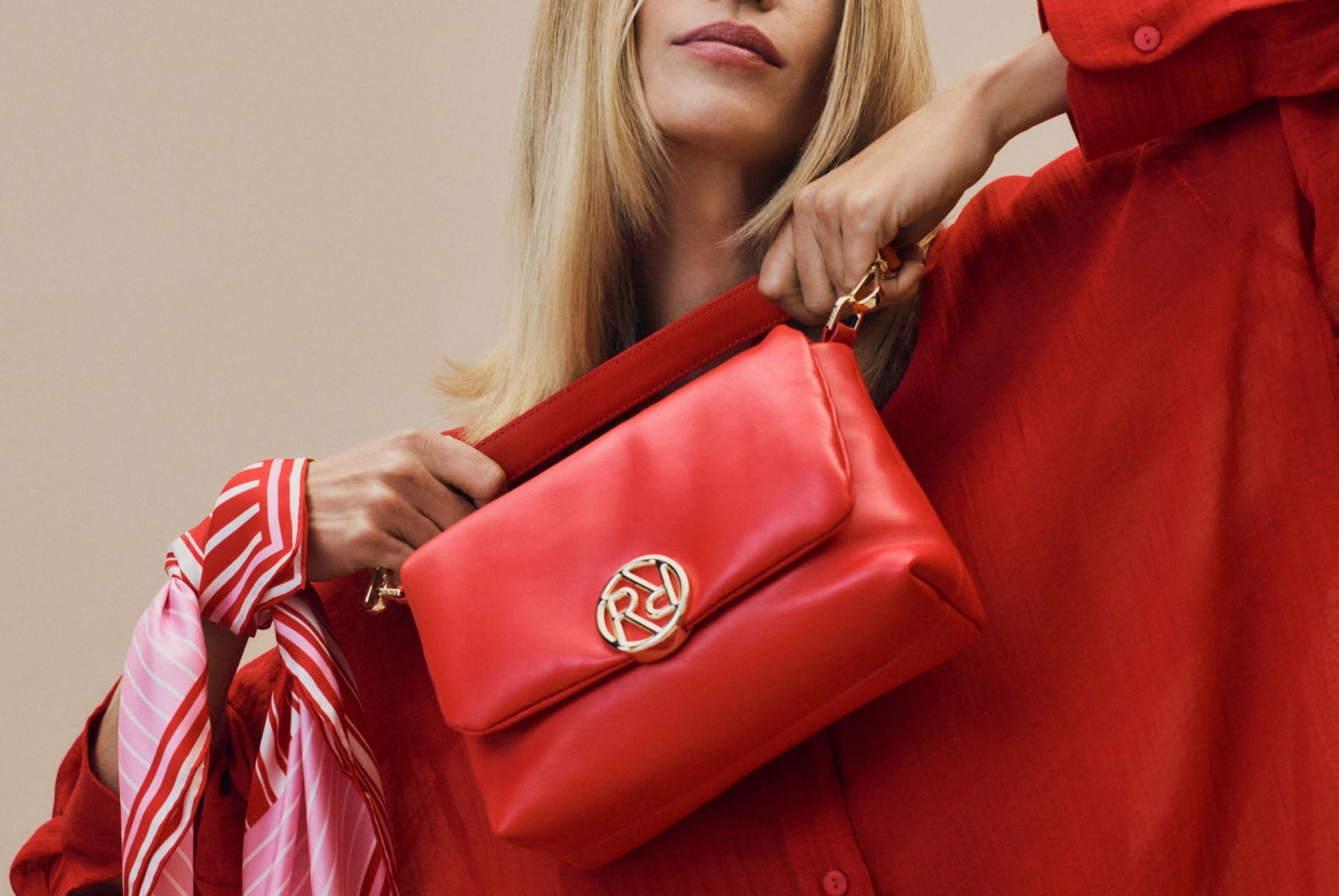 Eine blonde Dame in roten Kleidung hält eine rote Ledertasche in den Händen und hat um ihr Handgelenk ein rosa-rot gestreiftes Seidentuch.