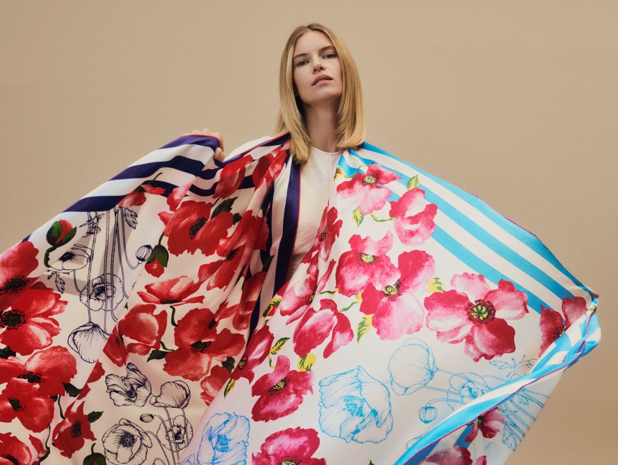 A blonde woman with shoulder-length hair is adorned with a large, colorful silk scarf featuring red poppies, blue stripes, and floral sketches.