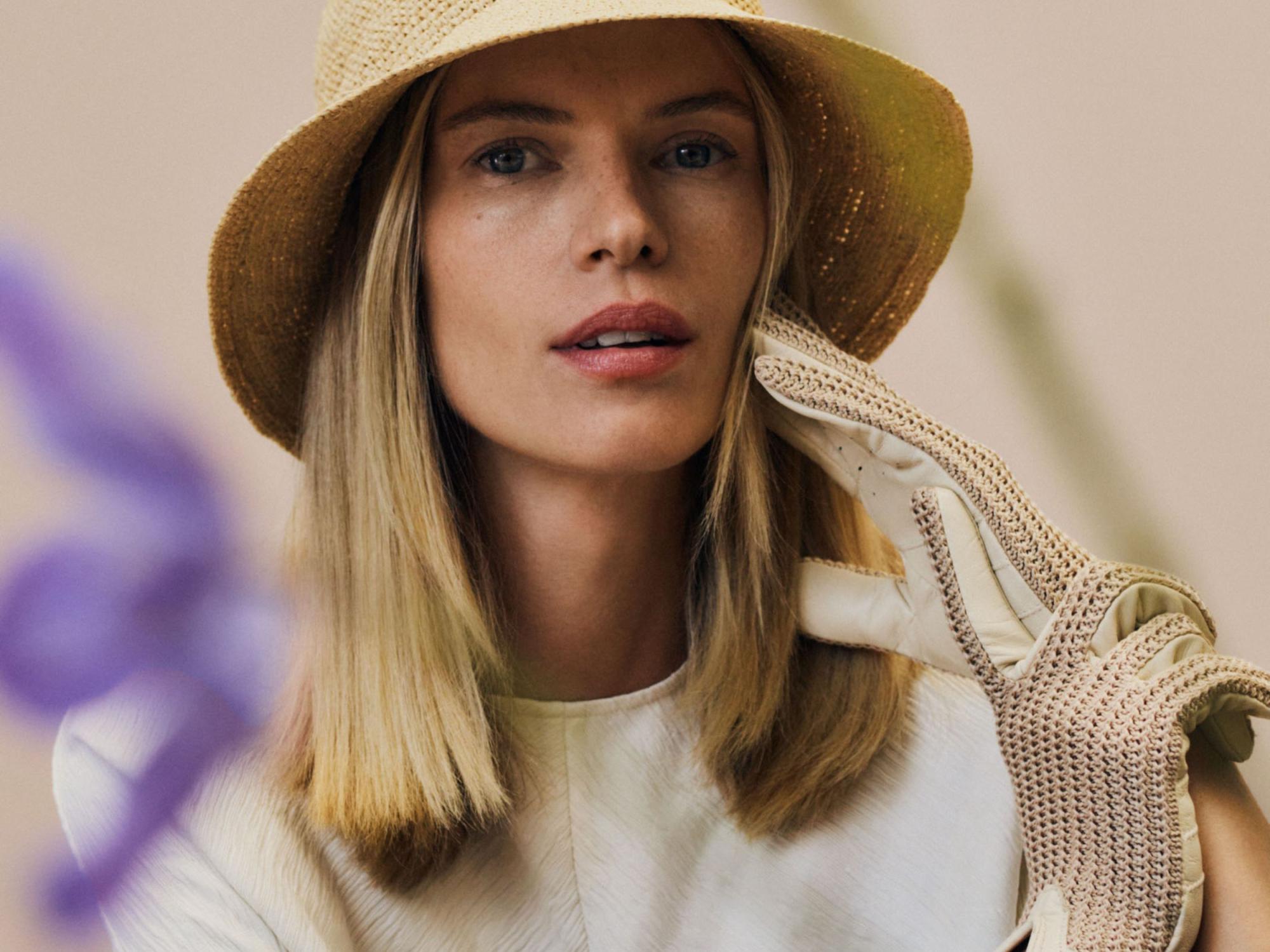 Eine blonde Frau trägt einen Papierstrohhut und geflochtene Handschuhe in beige.