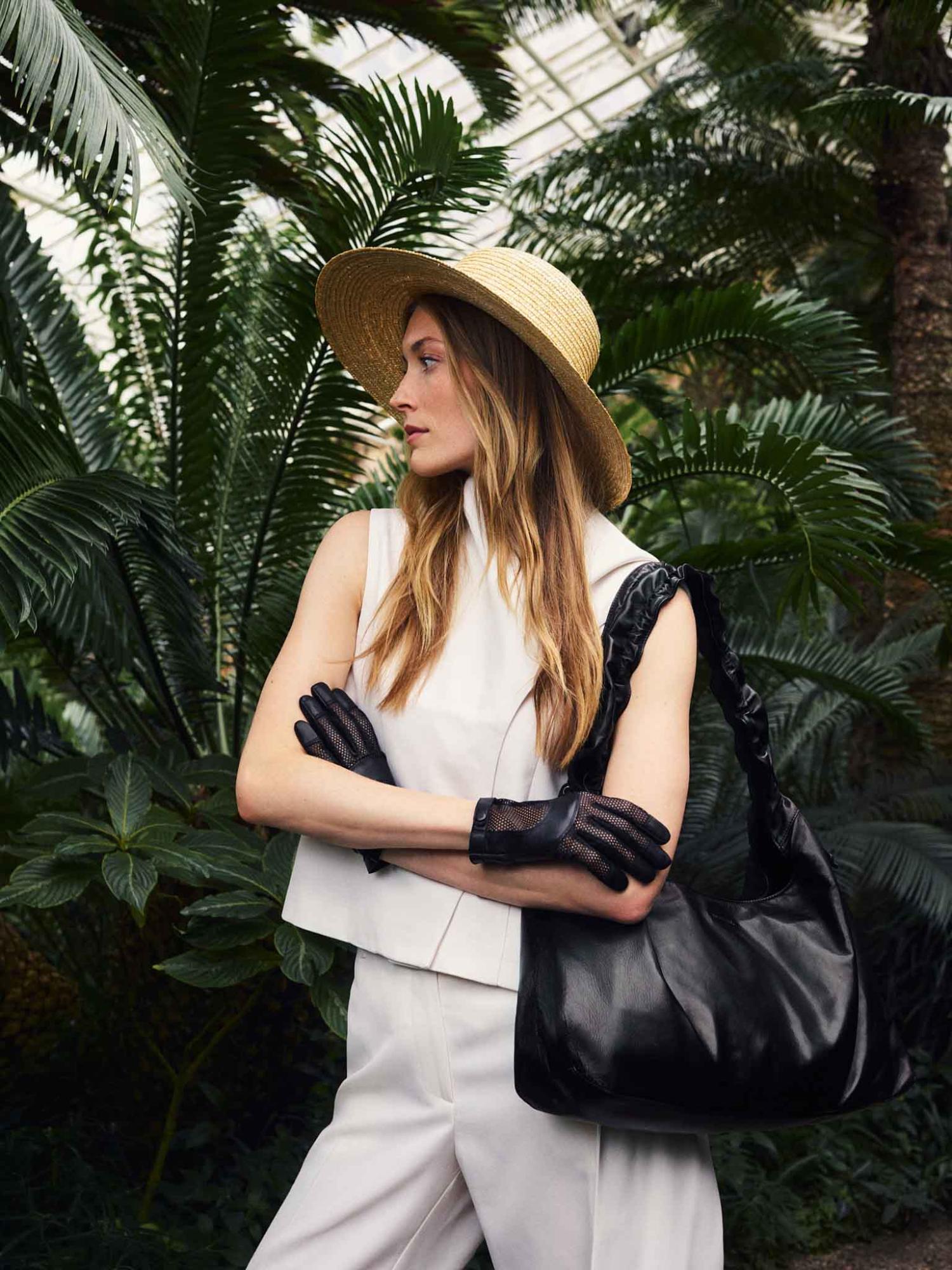 Frau im weißen Blazer mit schwarzen Lederhandschuhen hält eine schwarze ROECKL Lederhandtasche in einem botanischen Garten