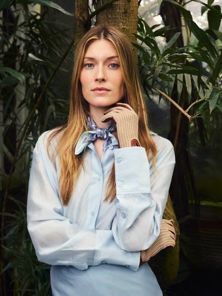 Frau mit hellblauer Bluse, Seidenschal und Häkelhandschuhen von ROECKL vor tropischer Pflanzenkulisse