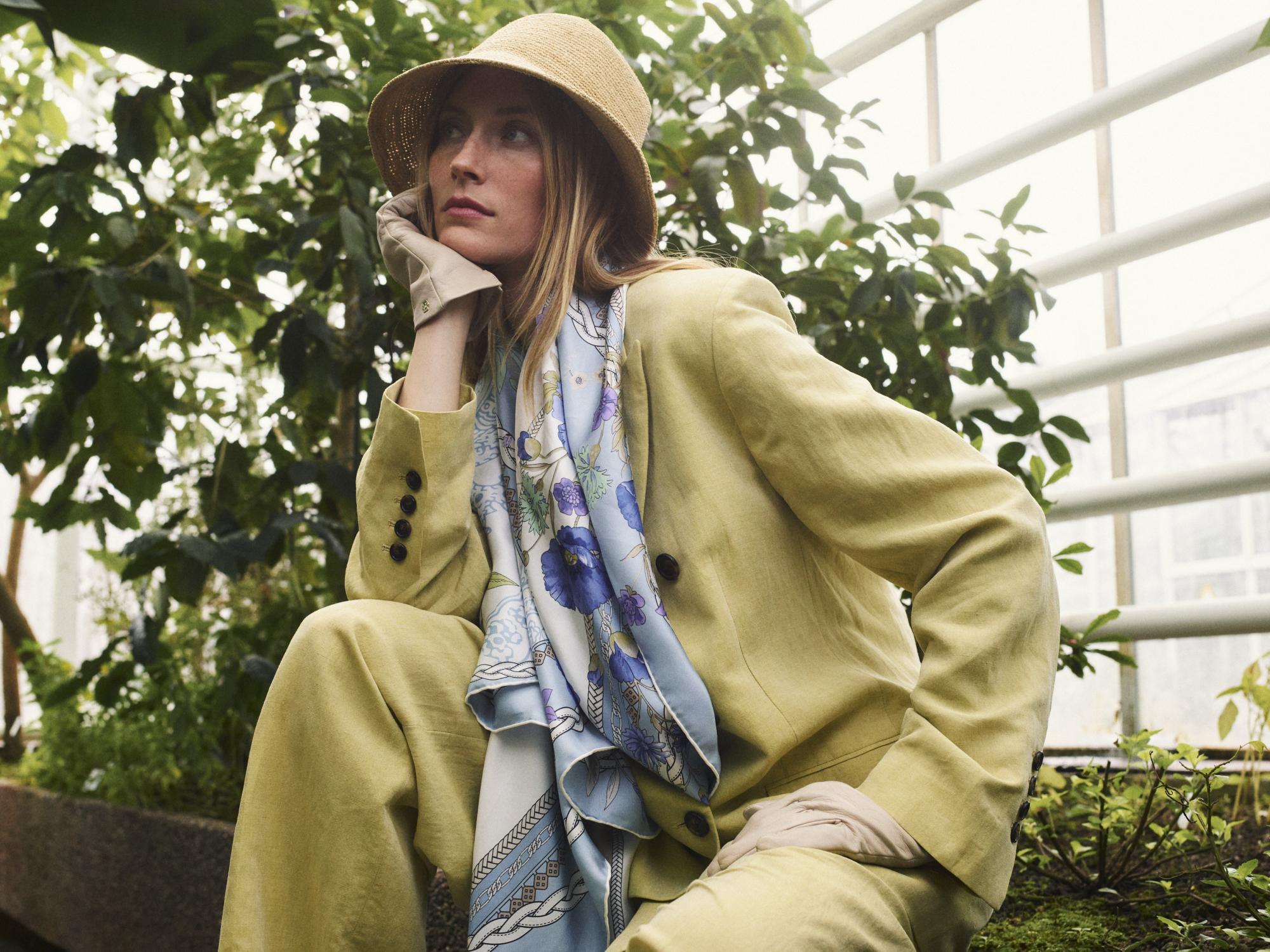 Eine blonde Dame in roten Kleidung hält eine rote Ledertasche in den Händen und hat um ihr Handgelenk ein rosa-rot gestreiftes Seidentuch.