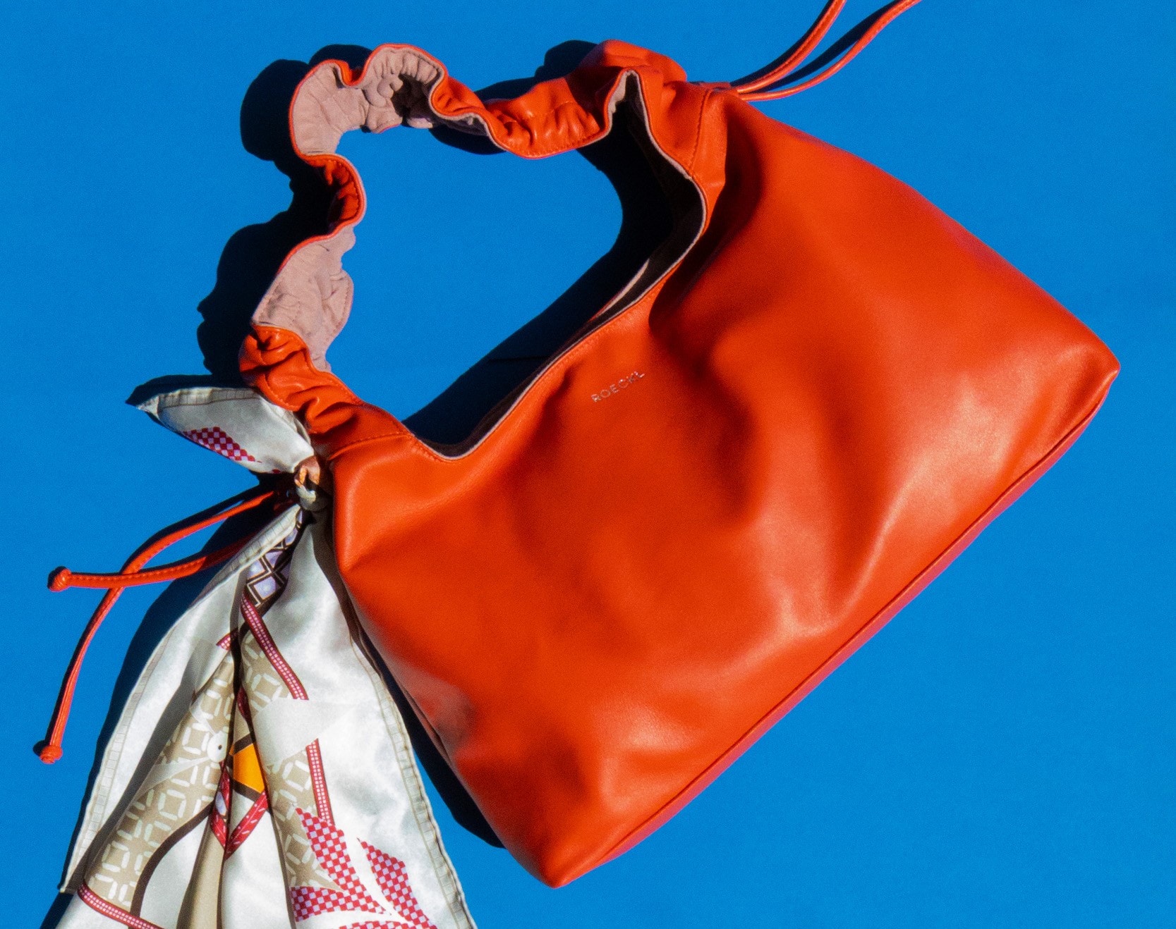 auffällige, farbintensive Tasche mit dekorativ gebundenem Tuch vor kontrastreichem Hintergrund.