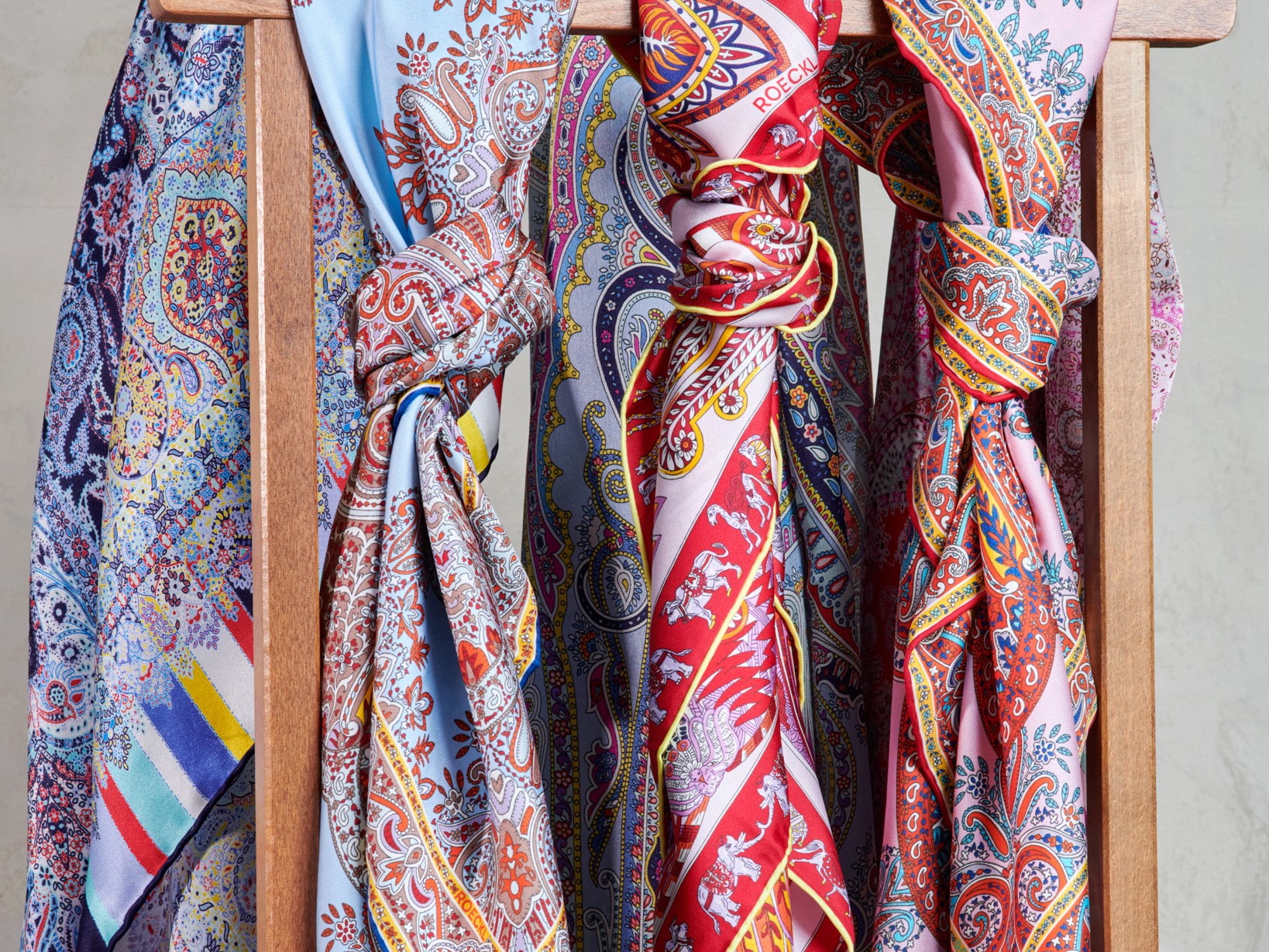 Roeckl Seidenschals mit detailreichen Paisley- und Ornamentmustern hängen dekorativ über einer Holzstruktur vor neutralem Hintergrund.