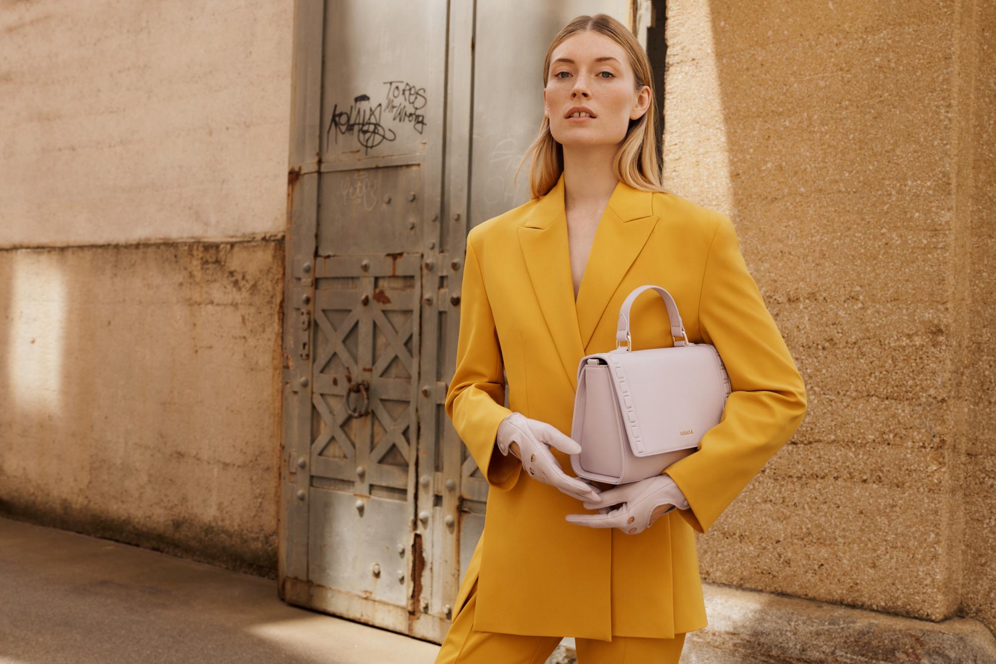 Frau in gelbem Hosenanzug trägt hellrosa ROECKL-Lederhandschuhe und passende strukturierte Handtasche – elegante Premium-Accessoires aus feinem Leder für einen modernen Business-Look.