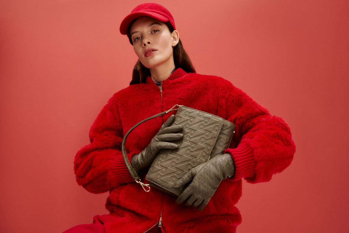 Frau in rotem Outfit mit taupefarbener ROECKL-Lederhandtasche und passenden strukturierten Lederhandschuhen – luxuriöses Accessoire-Set mit geprägtem Monogramm-Design.