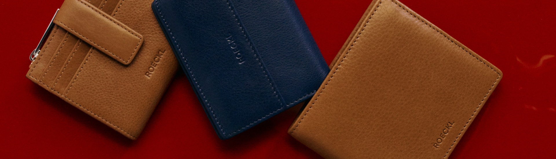 Mehrere Geldbörsen für Herren aus Leder in Braun und blauSeidentücher mit detaillierten Mustern, darunter florale und tierische Motive, in den Farben Rot, Blau, Weiß und Gelb, kunstvoll drapiert auf einer weißen Oberfläche.