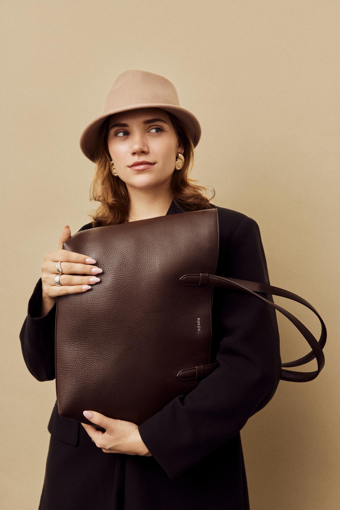 Blonde Dame trägt ein großes Seidentuch um die Schultern und hält eine braune Ledertasche in der Hand.