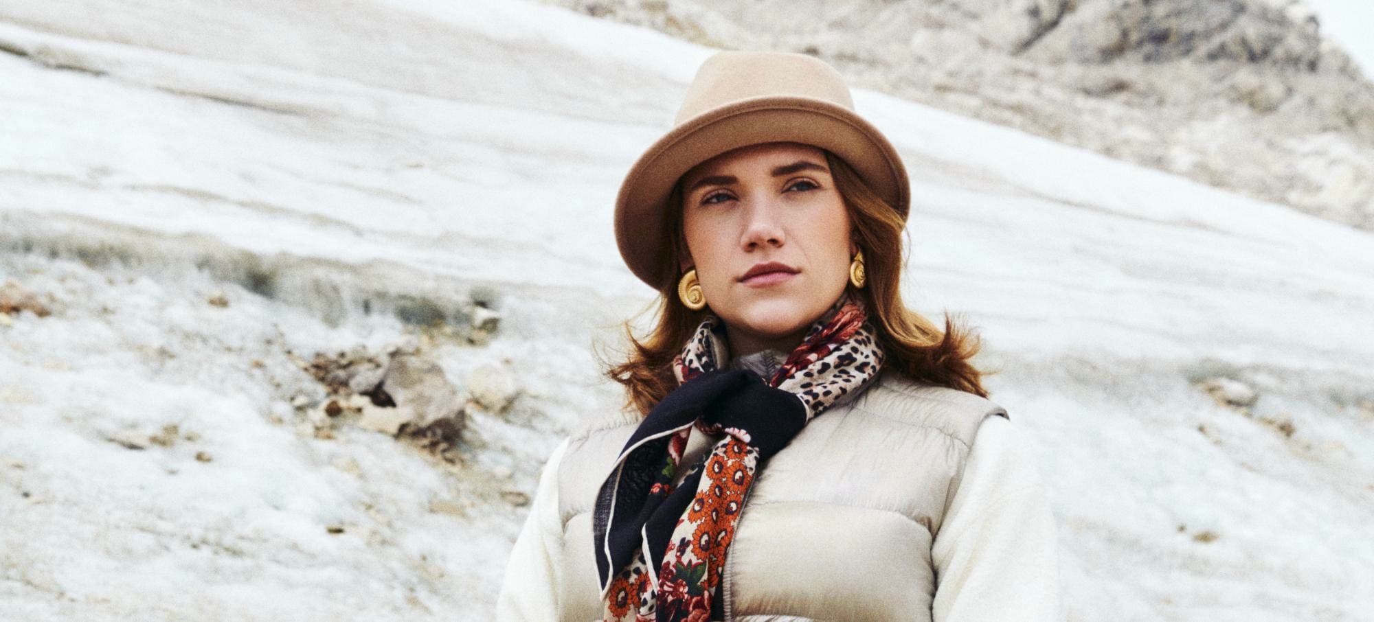 Zwei stilvoll gekleidete Damen stehen nebeneinander. Die Dame auf der rechten Seite hält eine beige Ledertasche, trägt Handschuhe im selben Ton und hat ein beiges Tuch über die Schulter drapiert. Die Dame auf der Linken Seite trägt einen Hut in Burgunder eine Handtasche in dem selben Ton.