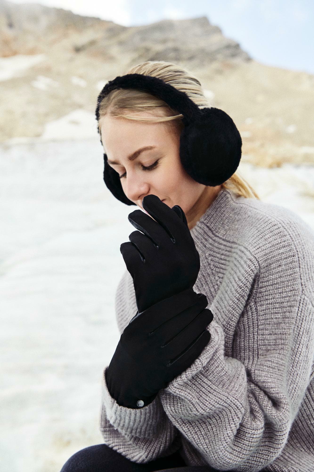 Frau in winterlicher Landschaft mit schwarzen ROECKL-Handschuhen, flauschigen Ohrwärmern und grauem Strickpullover – stilvolles, warmes Winteraccessoire-Outfit.