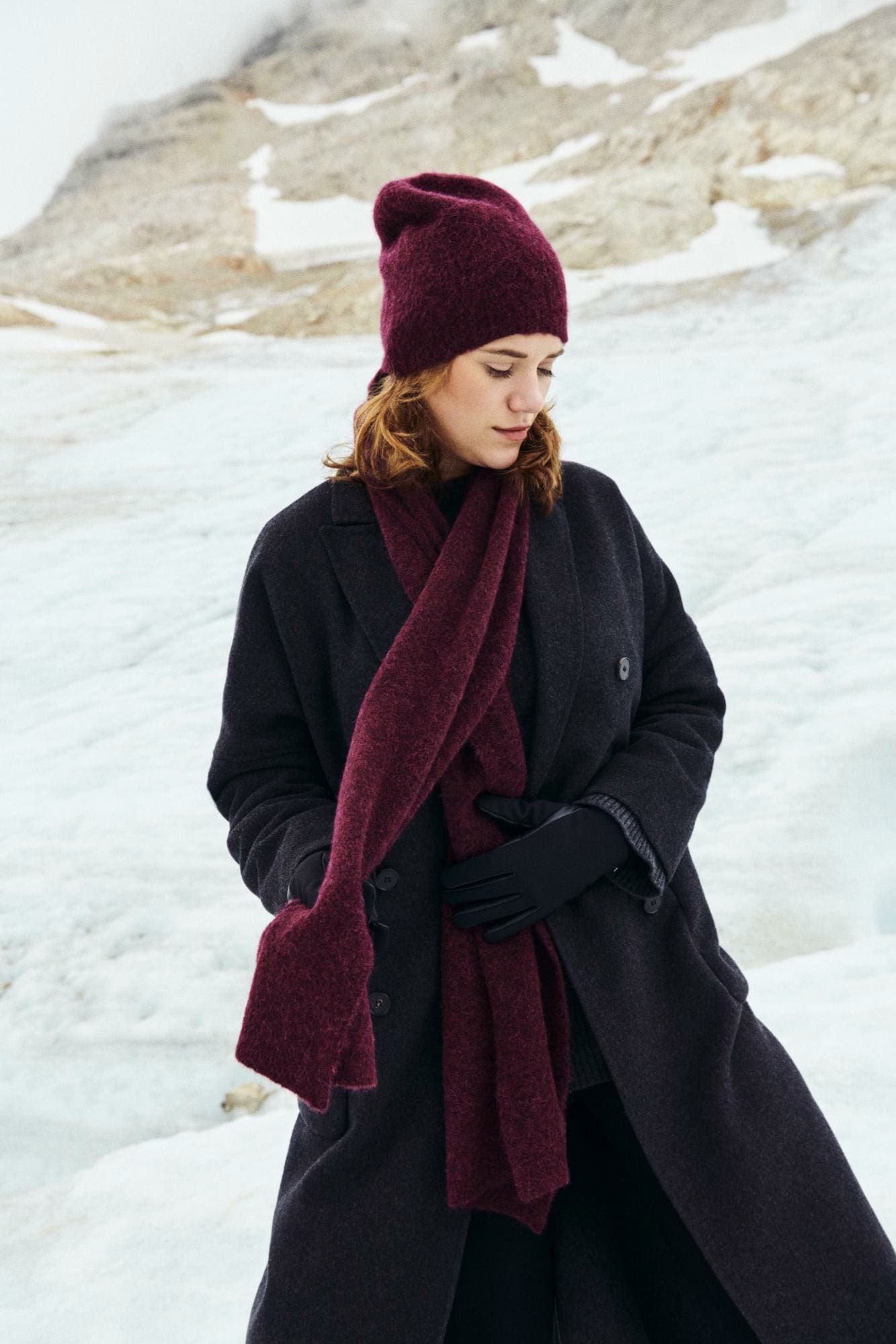 Frau trägt bordeauxrote Mütze und Schal aus weicher Wolle, kombiniert mit dunkelgrauem Mantel und schwarzen Handschuhen in winterlicher Berglandschaft – elegantes Winter-Outfit für Damen.