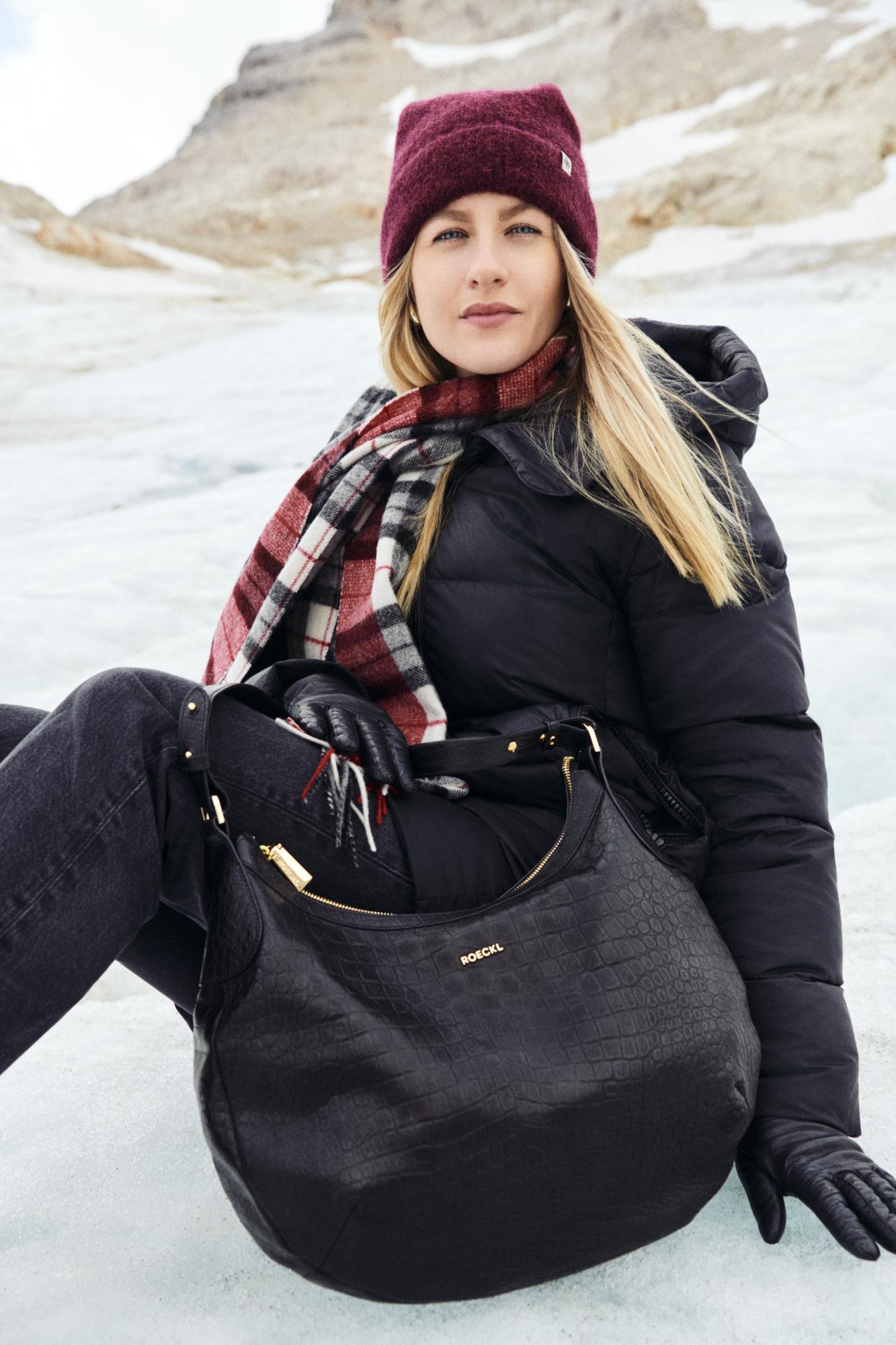 Frau in winterlicher Berglandschaft mit bordeauxfarbener Mütze, kariertem Schal, schwarzen Lederhandschuhen und großer schwarzer ROECKL-Tasche – stilvolles Winter-Outfit mit hochwertigen Accessoires.