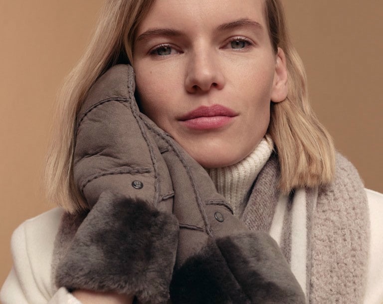 Frau in Winteroutfit mit grauen ROECKL-Lammfellhandschuhen, Wollschal und cremefarbenem Mantel – elegantes Accessoire für Wärme und Stil in der kalten Jahreszeit.