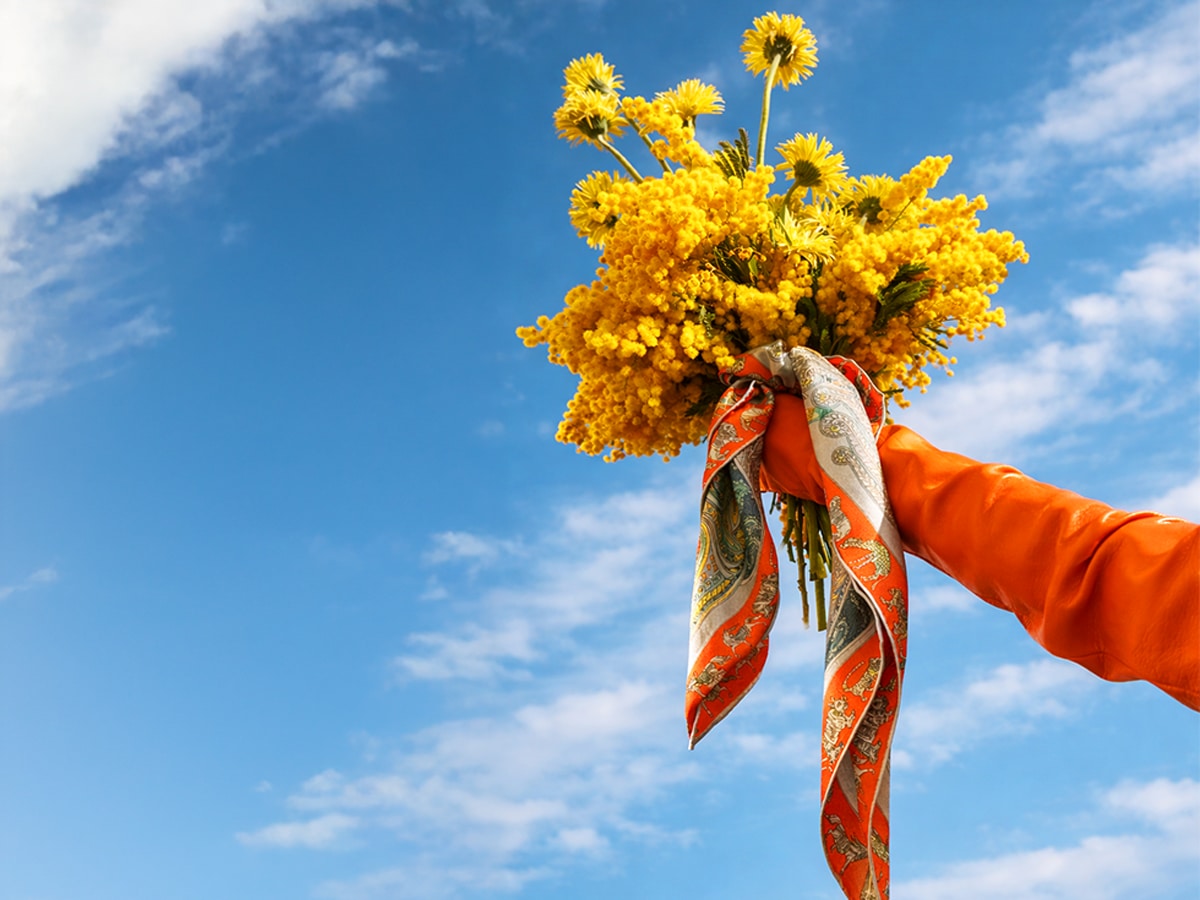 Eine blonde Frau mit schulterlangem Haar trägt ein großes, buntes Seidentuch mit roten Mohnblumen, blauen Streifen und floralen Skizzen.