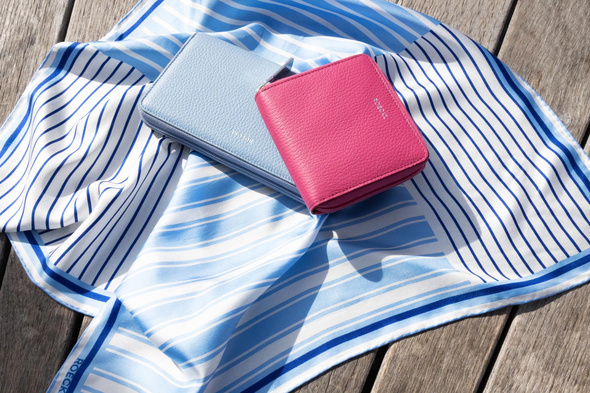Zwei ROECKL-Lederportemonnaies in Rosa und Hellblau auf einem blau-weiß gestreiften Seidenschal – stilvolle Accessoires in frischen Sommerfarben.