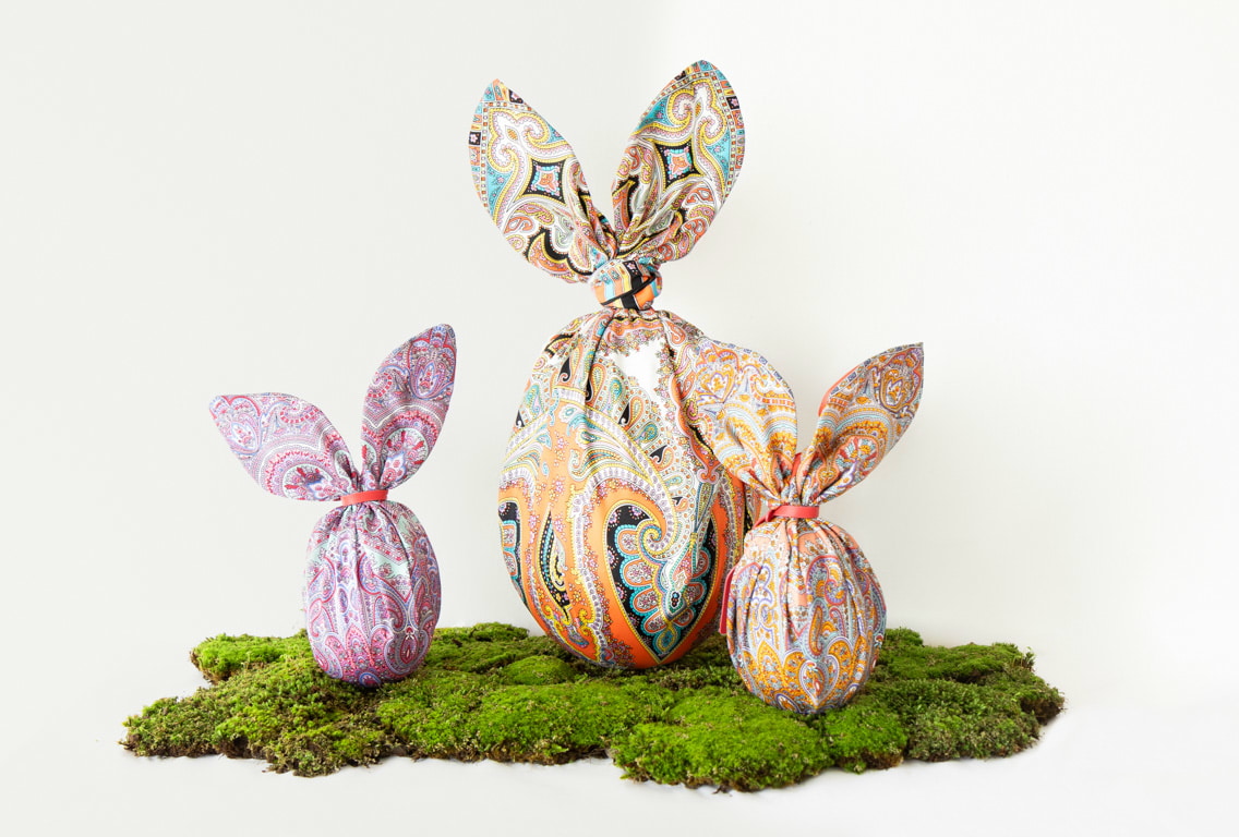 In der Mitte befinden sich drei Ostereier, die in ein buntes Tuch mit Hasenohren-Form eingewickelt sind. Die Szene ist hell und frühlingshaft gestaltet.