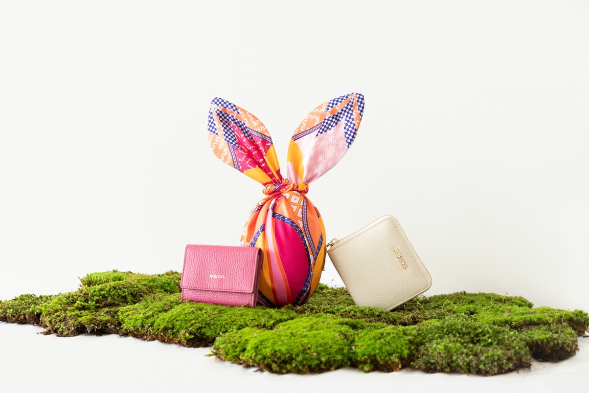 Two leather wallets in beige and pink are placed on green moss next to an Easter egg that has been wrapped in a silk cloth.