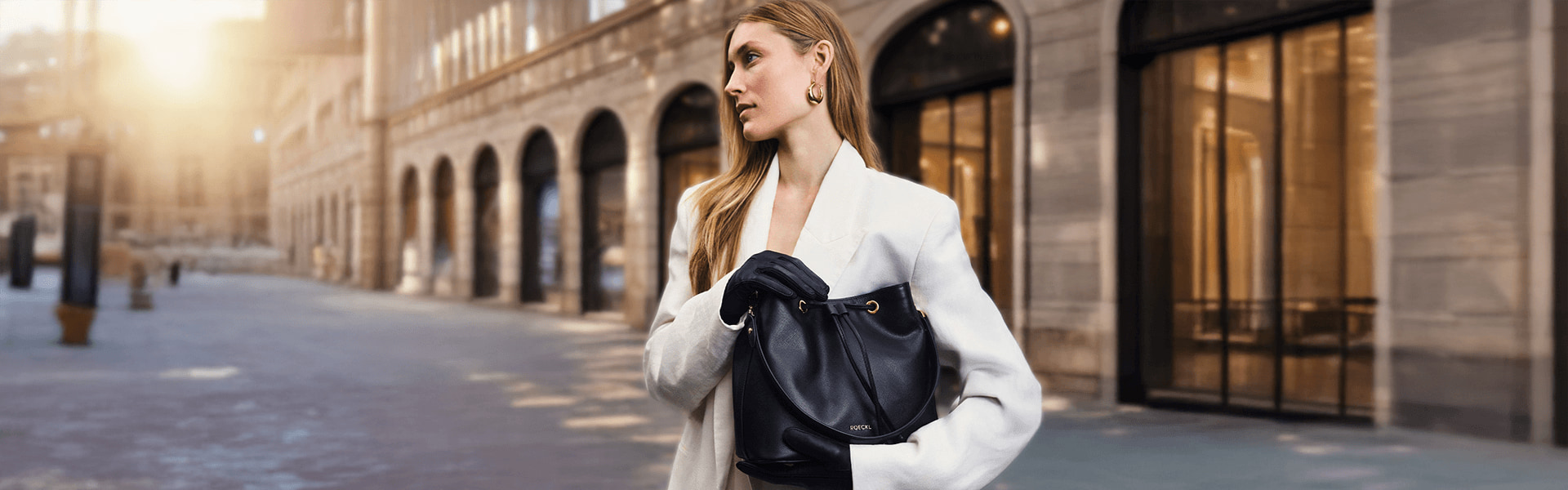Roeckl Lederhandtasche in Schwarz wird von einer eleganten Frau im weißen Blazer in einer urbanen Straßenszene mit historischer Architektur getragen.