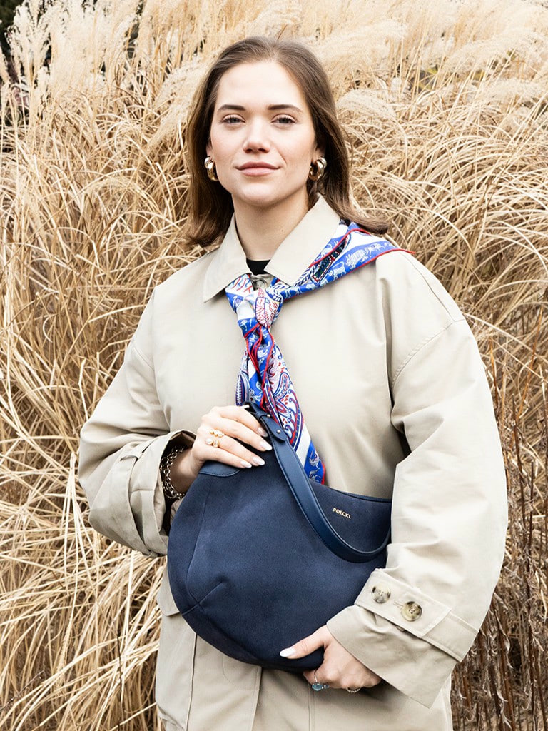 Frau mit beigefarbenem Trenchcoat, blauer ROECKL-Tasche aus Wildleder und gemustertem Seidentuch – klassischer Herbst-Look mit luxuriösen Akzenten.