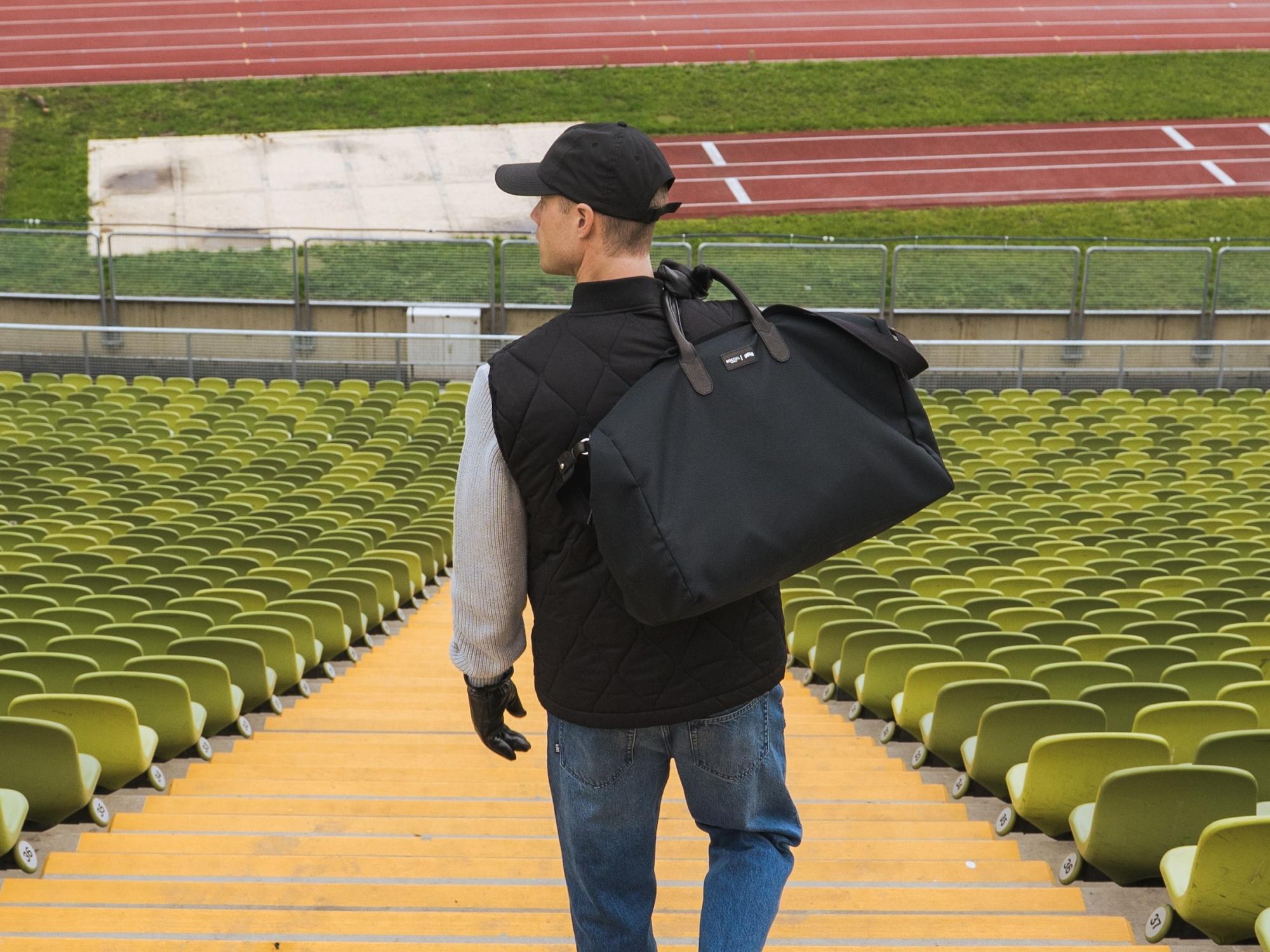 Roeckl schwarze Handschuhe werden von einem Mann getragen, der mit großer Tasche durch ein Stadion mit leeren Sitzreihen läuft.