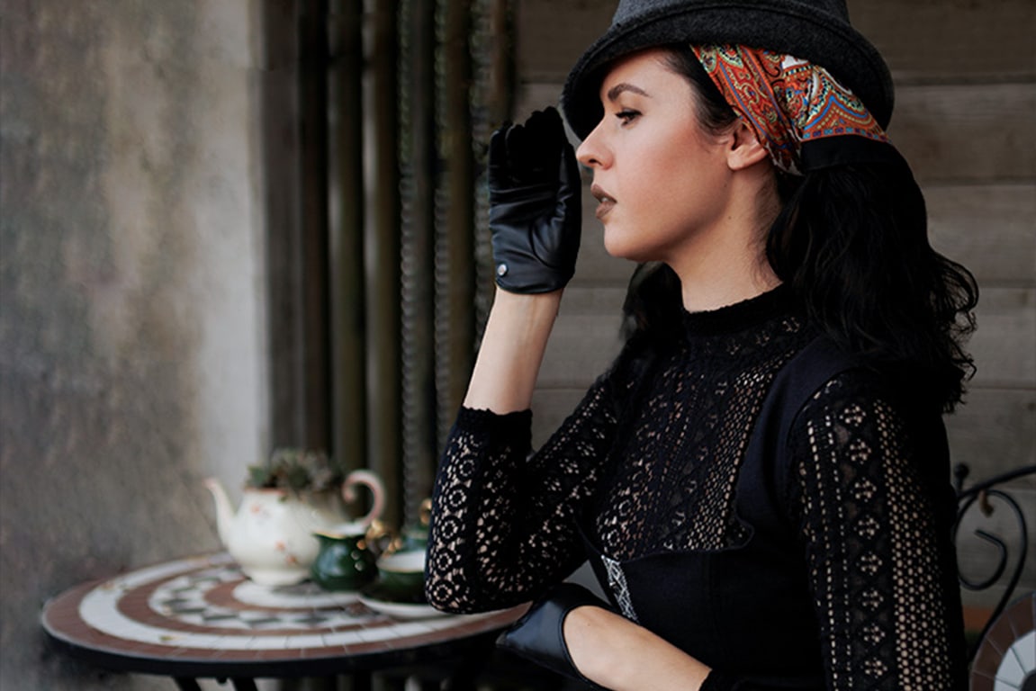 A woman with dark hair is wearing a dirndl, complemented by short black gloves, a gray felt hat, and a patterned silk scarf.