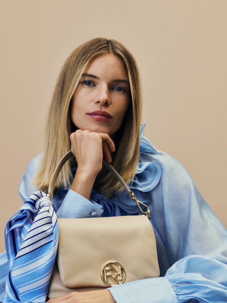 Eine blonde Dame in blauer Kleidung trägt eine beige Ledertasche und ein kleines gestreiftes Tuch in blau, dass um den Griff gebunden ist.